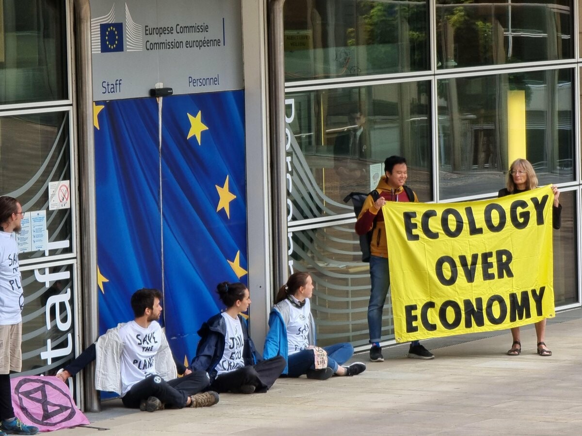 6 people block the entrance to the european commission. some are glued together. 2 hold a banner reading "Ecology over economy". the 4 others have messages on their shirts. "Save the planet", more messages are not readable.