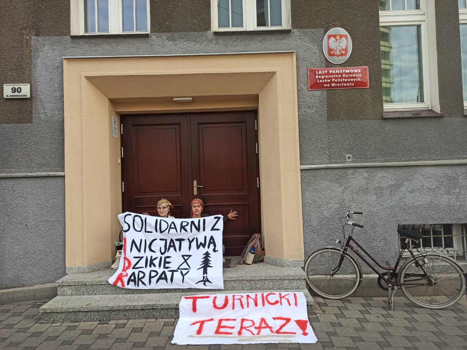 Osoby aktywistyczne przyklejone do drzwi, trzymający baner "Solidarni z Inicjatywą Dzikie Karpaty". Obok leży baner "Turnicki teraz!"