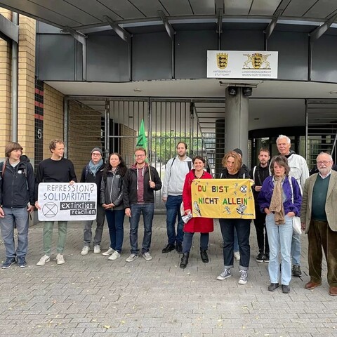 13 Menschen stehen vor dem Eingang des Amtsgericht Stuttgart.
Auf einen Demoschild steht:
Repression?
Solidarität!
Extinction Rebellion"
Auf den 2. gelben Schild steht in bunten Buchstaben:
"Du bist nicht allein"