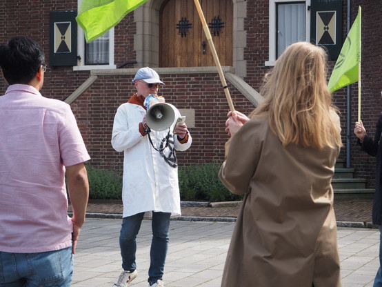 Een close-up van een man in een witte wetenschappersjas die in een megafoon praat. Op de voorgrond mensen die om de wetenschapper heen staan, met XR vlaggen.