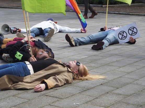 Close-up van een aantal mensen die voor dood liggen op een grond met tegels. Sommigen houden XR vlaggen of borden omhoog