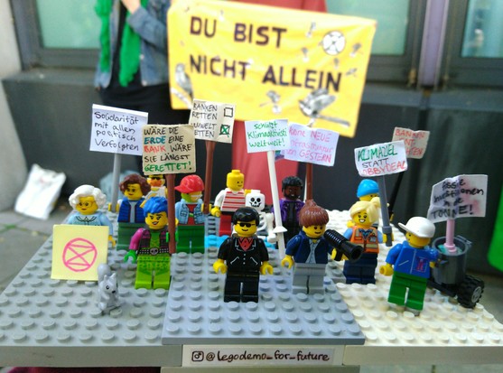 Lego bzw Klemmbausteinfiguren stehen auf grauen Platten beisammen und halten kleine Schildchen auf denen z.B  steht:
XR-Logo (pink)
"Wäre die Erde eine Bank, wäre sie längst gerettet."
"Keine neue Infrastruktur von gestern!"
"Banken entmachten"
"fossile Investitionen in die Tonne"