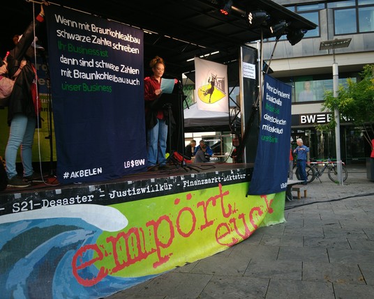 Einen Bühne steht auf den kleinen Schlossplatz Stuttgart vor einen großen LBBW-Gebäude. An der Bühnenkante hängt ein grünes Banner bis zum Boden mit einer türkisen Welle abgebildet, in roter Schrift steht groß: "empört euch" Darüber  ".. - S21-Desaster - Justizwillkür - Finanzmarkt-Dikatatur" 
Auf der Bühne werden rechts und links in dunkelblau 2 Banner gehalten auf den steht links: "Wenn mit Braunkohleabbau schwarze Zahlen schreiben Ihr Business ist, dann sind schwarze Zahlen mit Braunkohletaabbau auch unser Business
LBBW" und rechts:
"Wenn Kohle machen mit Braunkohleabbau Ihr Business ist
dann ist Kohle machen mit Braunkohle auch unser Business.
LBBW"
Zwischen den Banner steht eine Rednerin und liest einen Text vor.
Im Hintergrund ist ein LBBW-Gebäude und teilweise der Schriftzug "BW Bank" zu sehen.