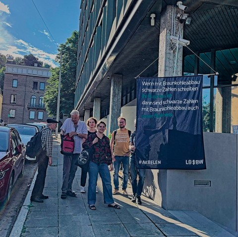 6 Menschen stehen solidarisch vor dem Landgericht und haben ein großes dunkelblaues Banner dabei auf den steht:
"Wenn mit Braunkohleabbau schwarze Zahlen schreiben ihr Business ist
dann sind schwarze Zahlen mit Braunkohleabbau auch unser Business

#Akbelen   LBBW"
